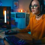 a woman playing Manor Lords on her PC in a dimly lit room