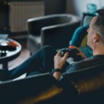 An adult man sitting on a sofa and holding a joystick while playing Dying Light: The Beast on console