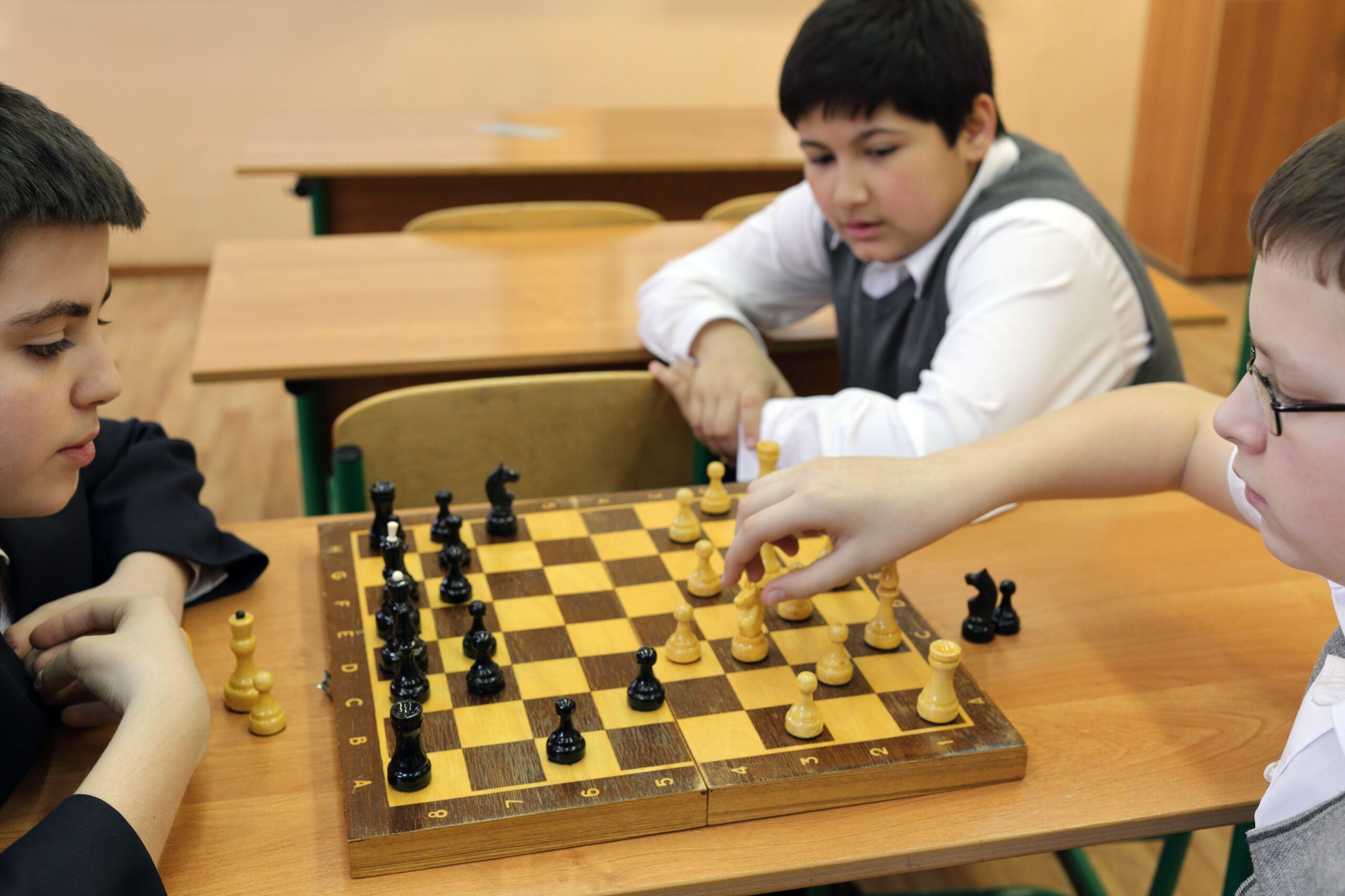 three children sitting in a classroom with two playing Chess